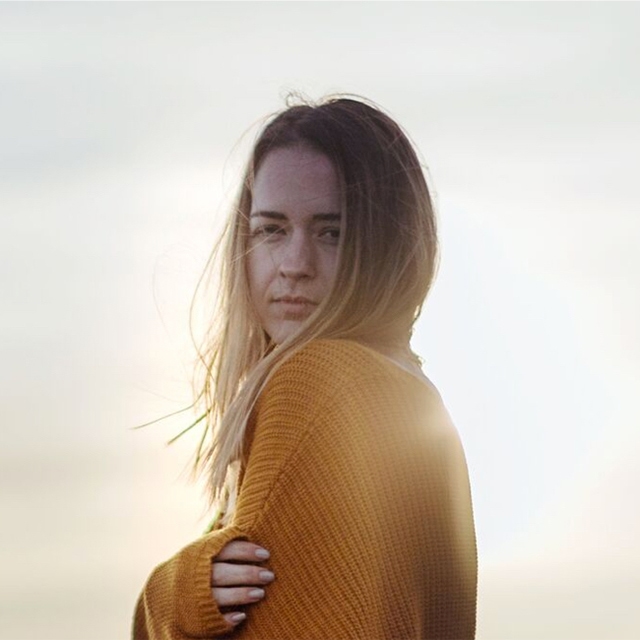 Woman in a mustard sweater stands against a soft, bright background.